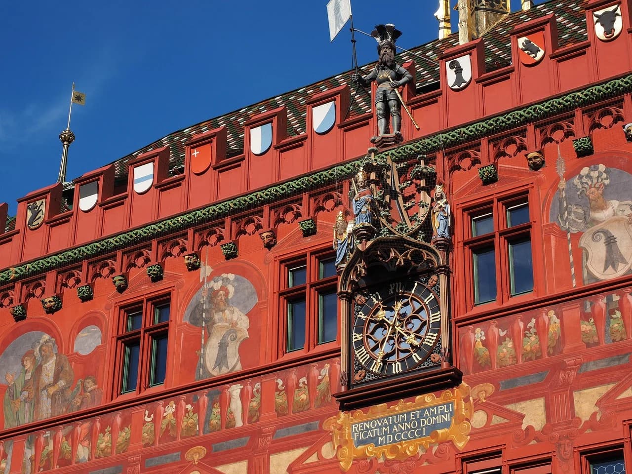 A close-up of the ornate, red facade of the Basel Town Hall, with its astronomical clock and intricate statues.