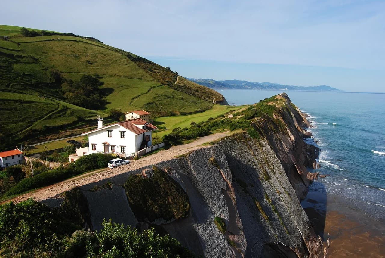 A white house is perched on the edge of a dramatic cliff, with the rugged coastline and the blue ocean in the background.