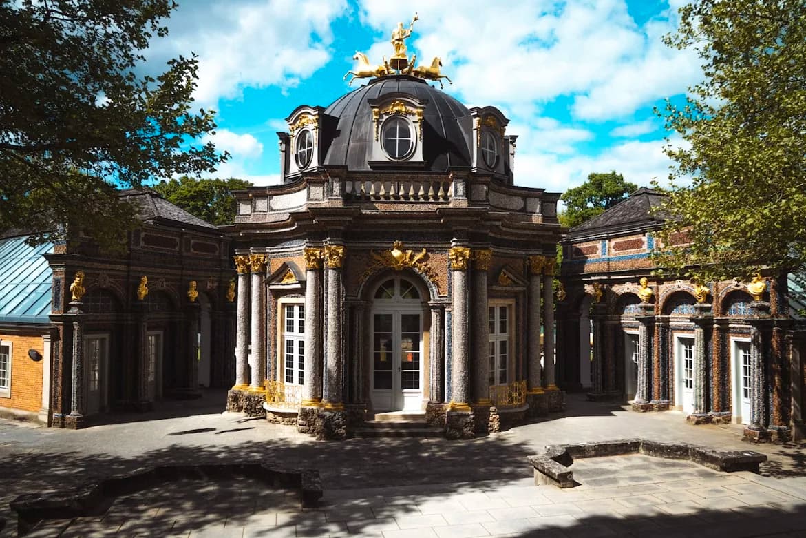A beautiful, ornate palace building with a central dome and large columns is surrounded by lush green trees and manicured gardens.