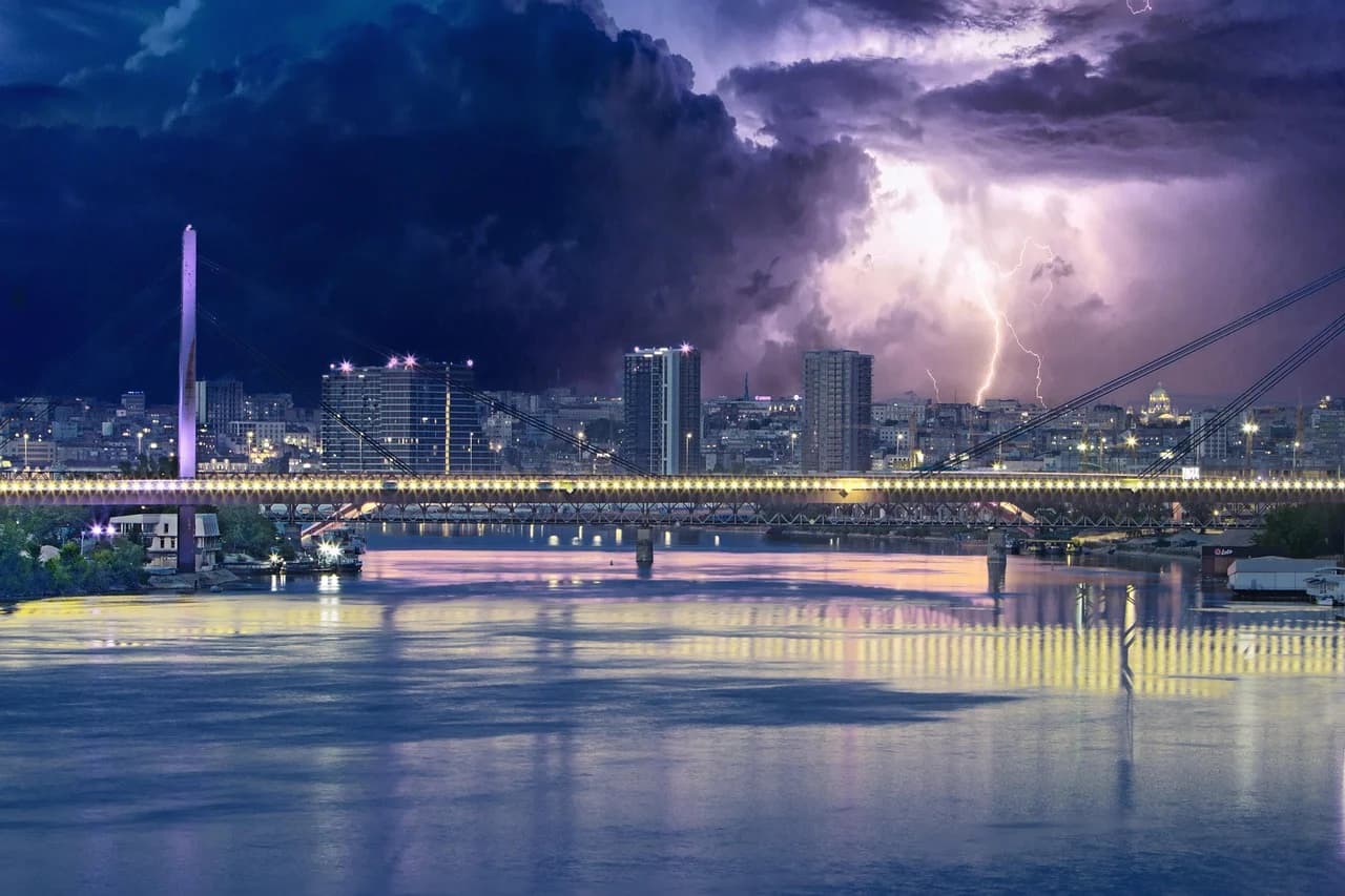 A modern bridge is illuminated at night, spanning a river with the city lights and a lightning storm in the background.