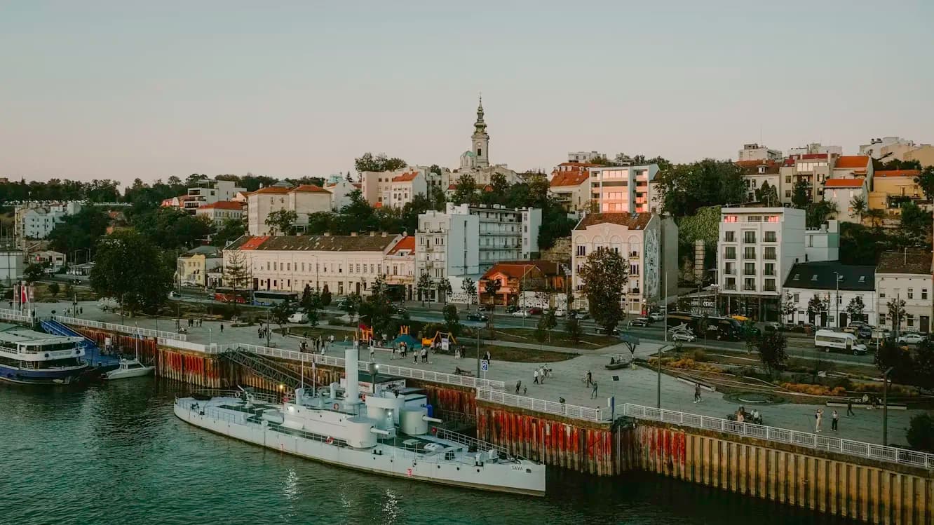 A large, historic military ship is moored at a pier on the river, with the residential buildings of Belgrade's Zemun district in the background.