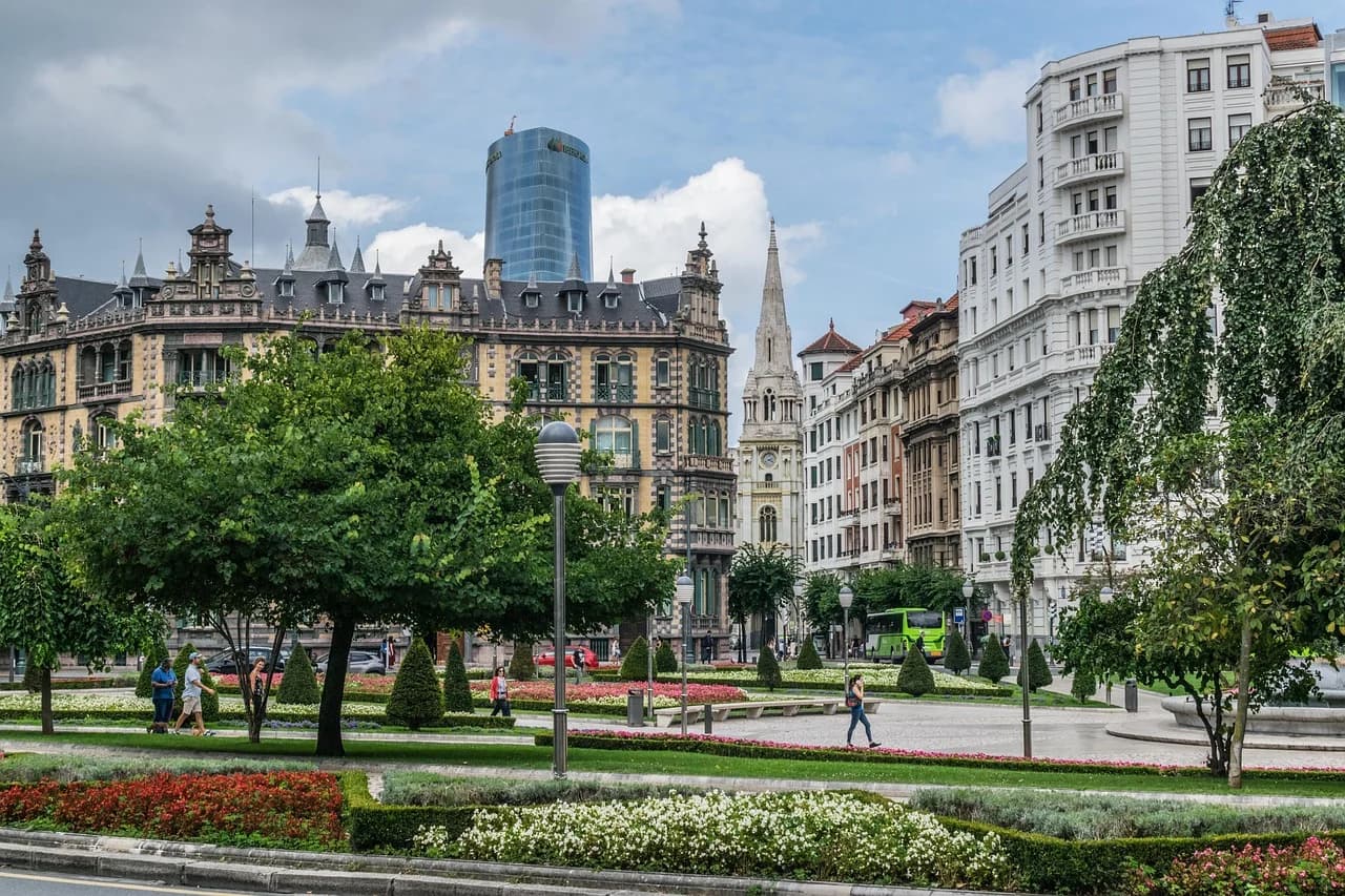 The bustling city street is filled with people, green trees, and historic buildings, with a view of a church steeple and a modern skyscraper.
