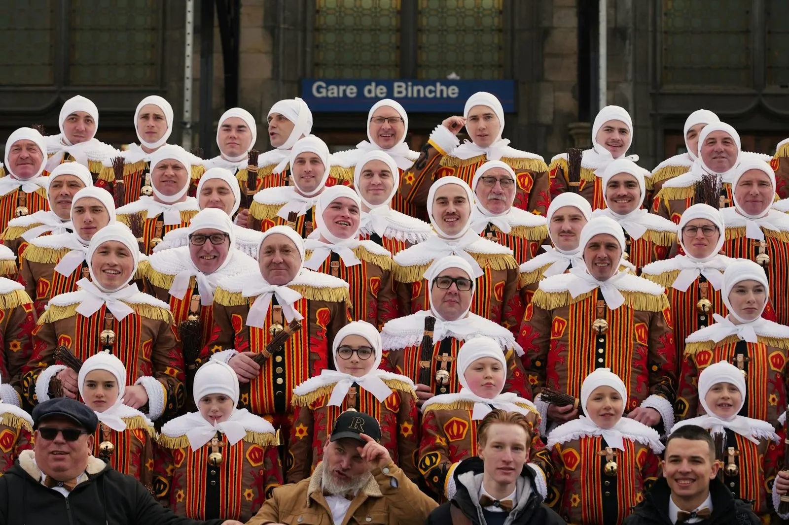 A large group of people in traditional Carnival of Binche costumes stand in front of a sign for the "Gare de Binche".