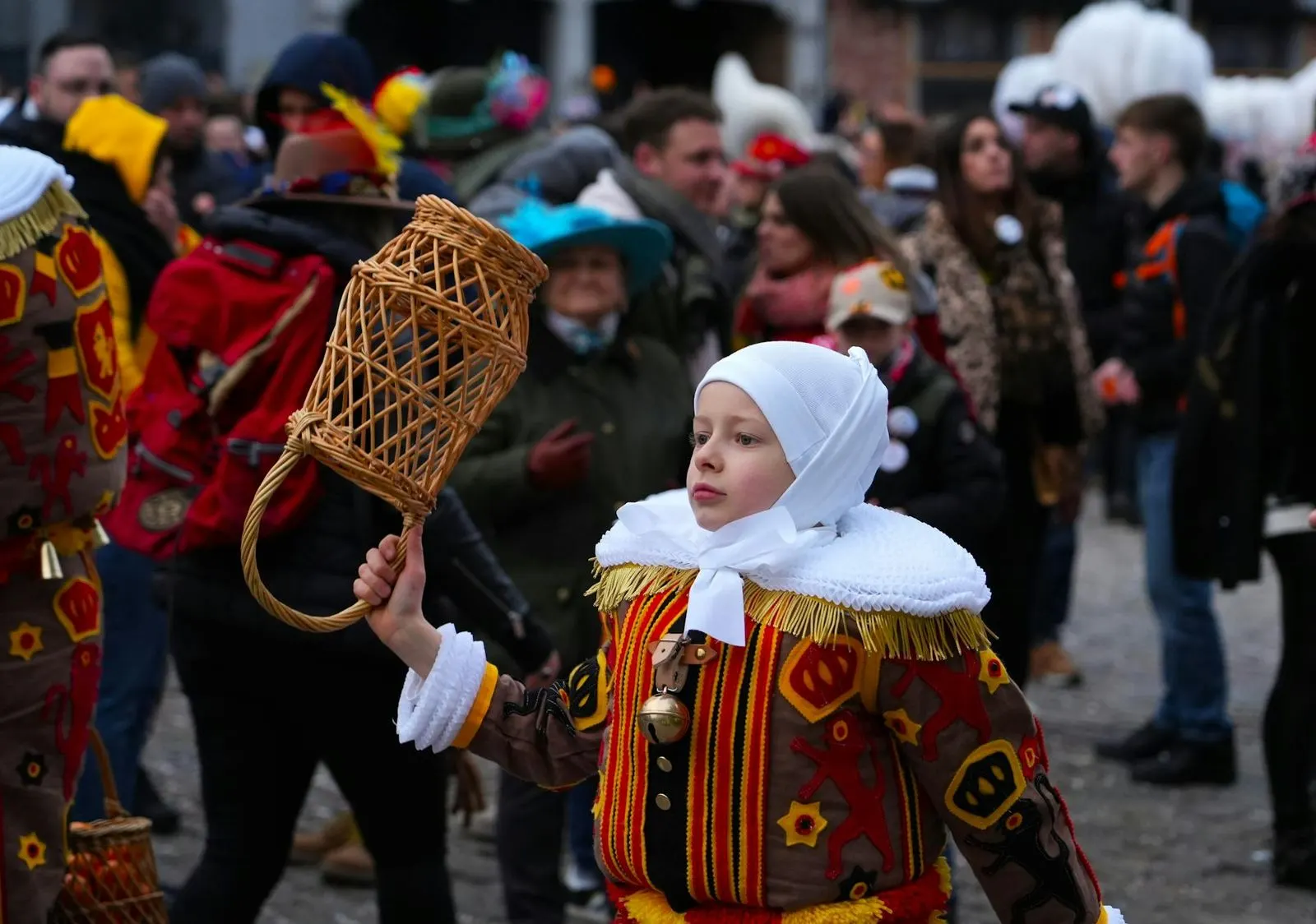 A young boy in a traditional Carnival of Binche costume holds a basket, with a crowd of people in the background.