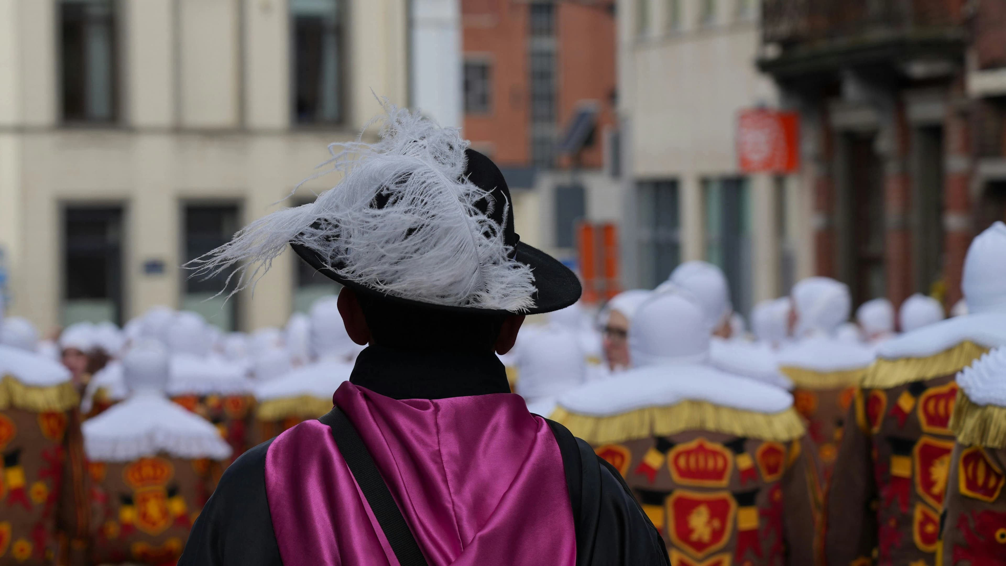 The back of a person in a traditional Carnival of Binche costume with a black feathered hat and a pink sash is seen in a crowd.