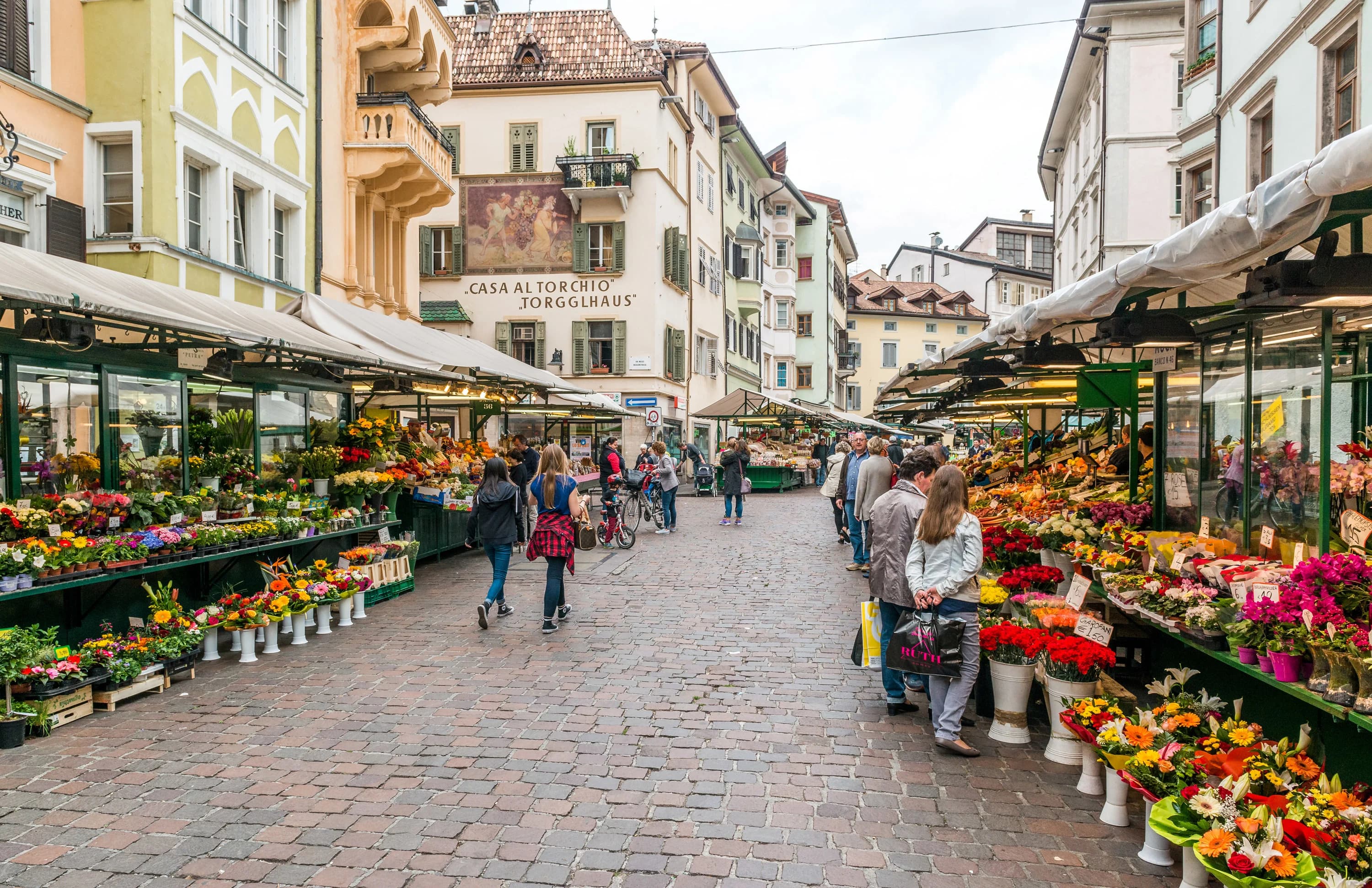 A vibrant open-air market in a cobblestone square is filled with stalls selling flowers and local produce.