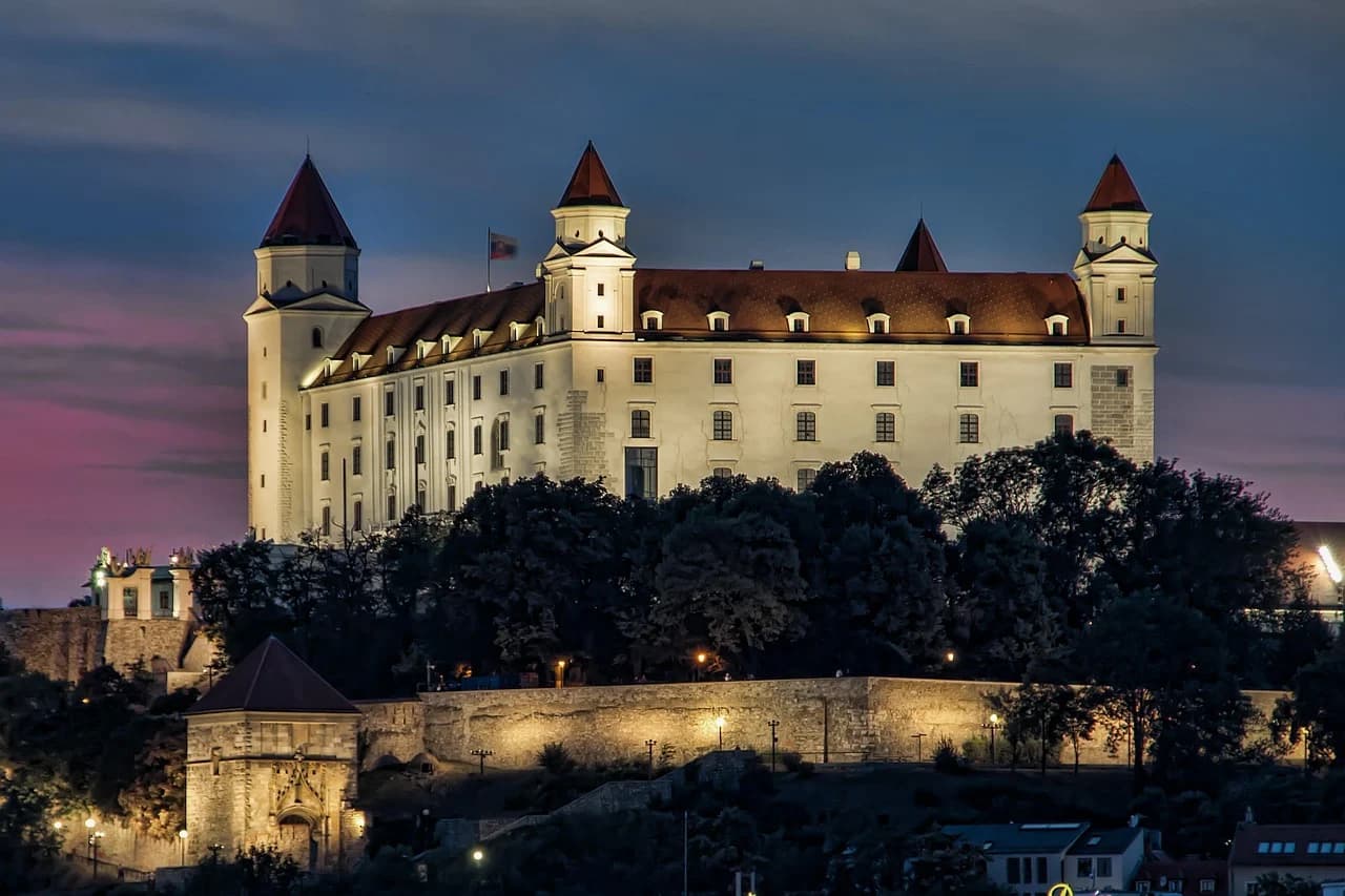 The majestic Bratislava Castle is illuminated at night, with the light reflecting on the surrounding greenery and a dark sky in the background.