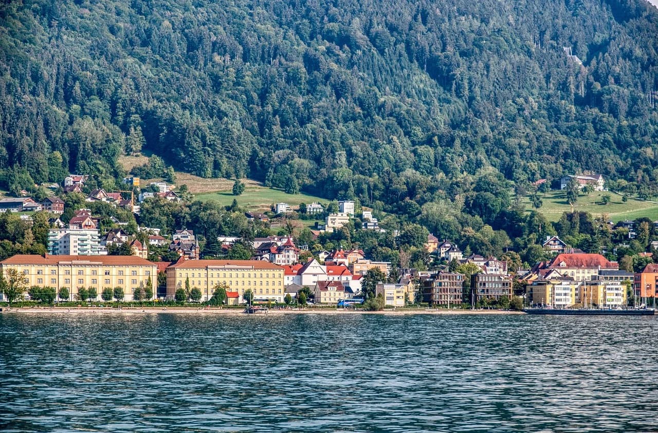 The city of Bregenz is nestled at the base of a forested mountain, with its buildings lining the shore of Lake Constance.