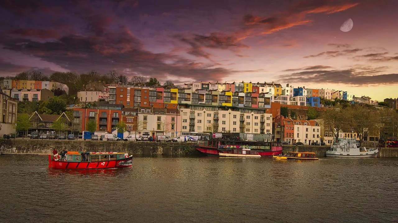 A small, red boat with a shark mouth painted on its front moves down the Avon River, with colorful houses and buildings lining the banks at sunset.