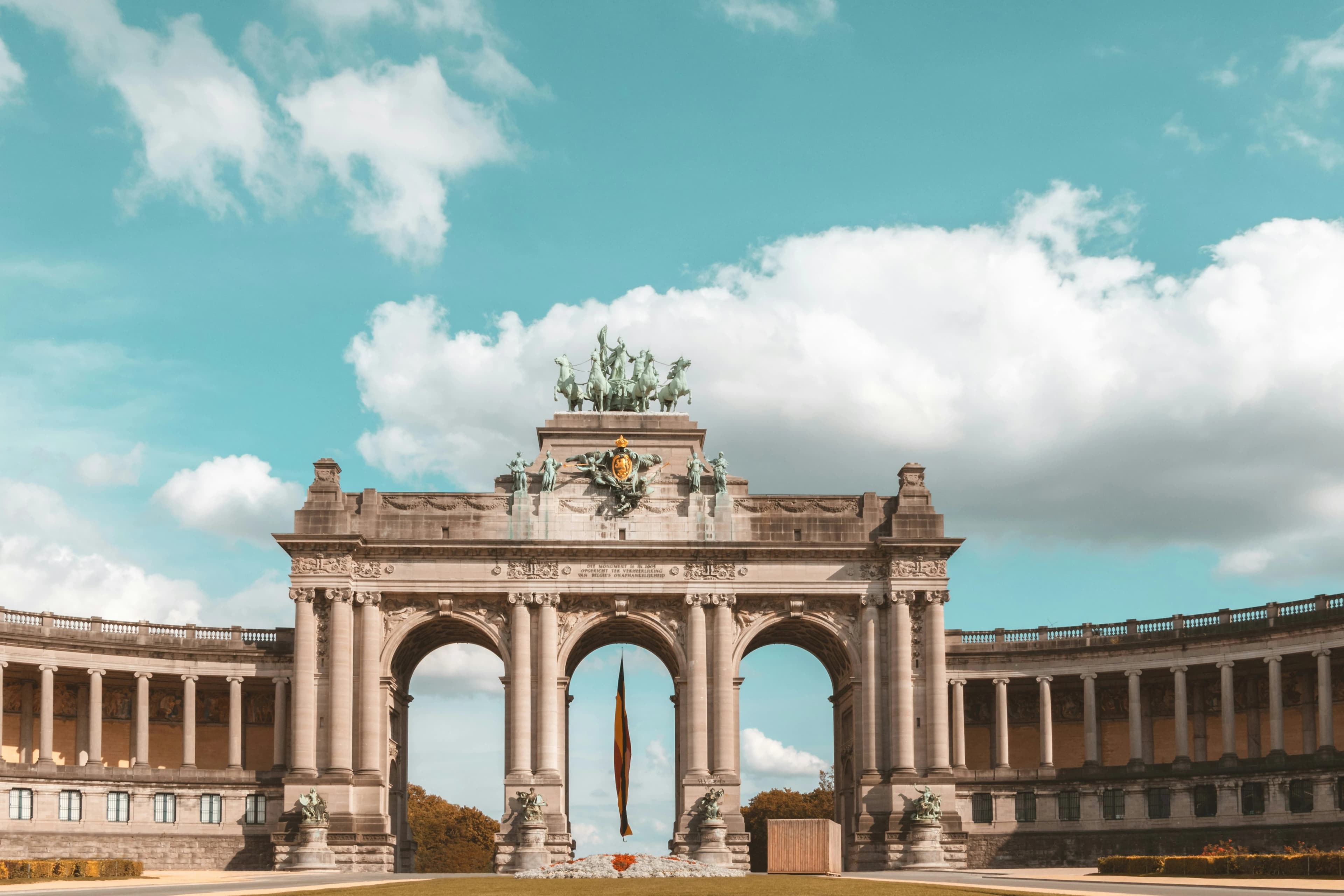 The Triumphal Arch of the Cinquantenaire, a monumental arch with three arches and a bronze chariot sculpture, stands in a park.