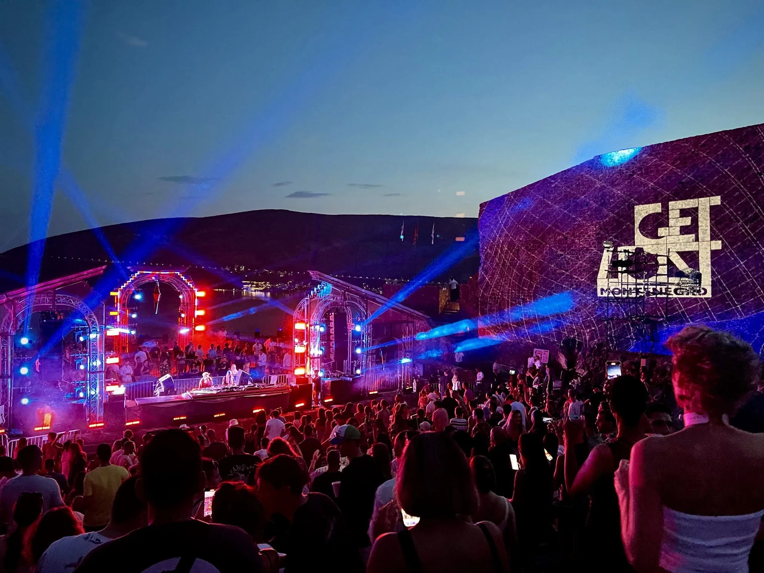 A music festival is in full swing on a stage overlooking the sea, with a large crowd of people dancing and enjoying the night.