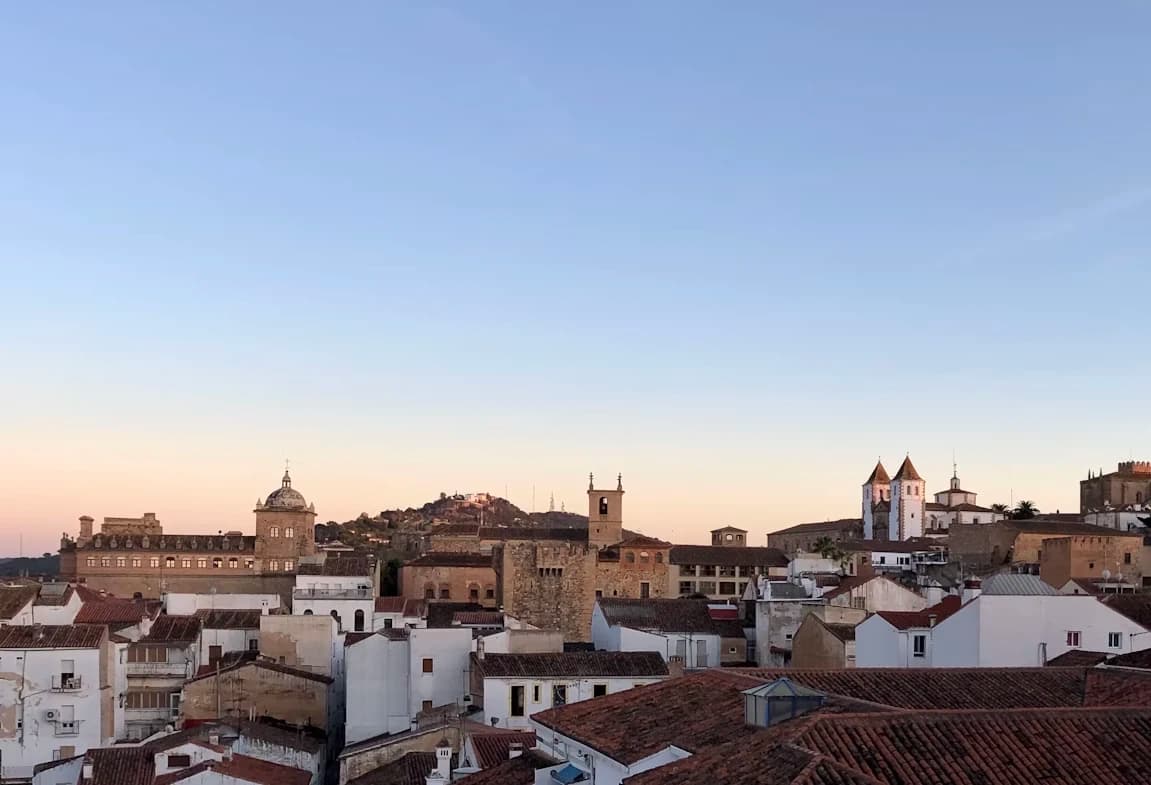 The historic city of Cáceres is a beautiful mosaic of old buildings and rooftops, with a fortress on a distant hill at sunset.