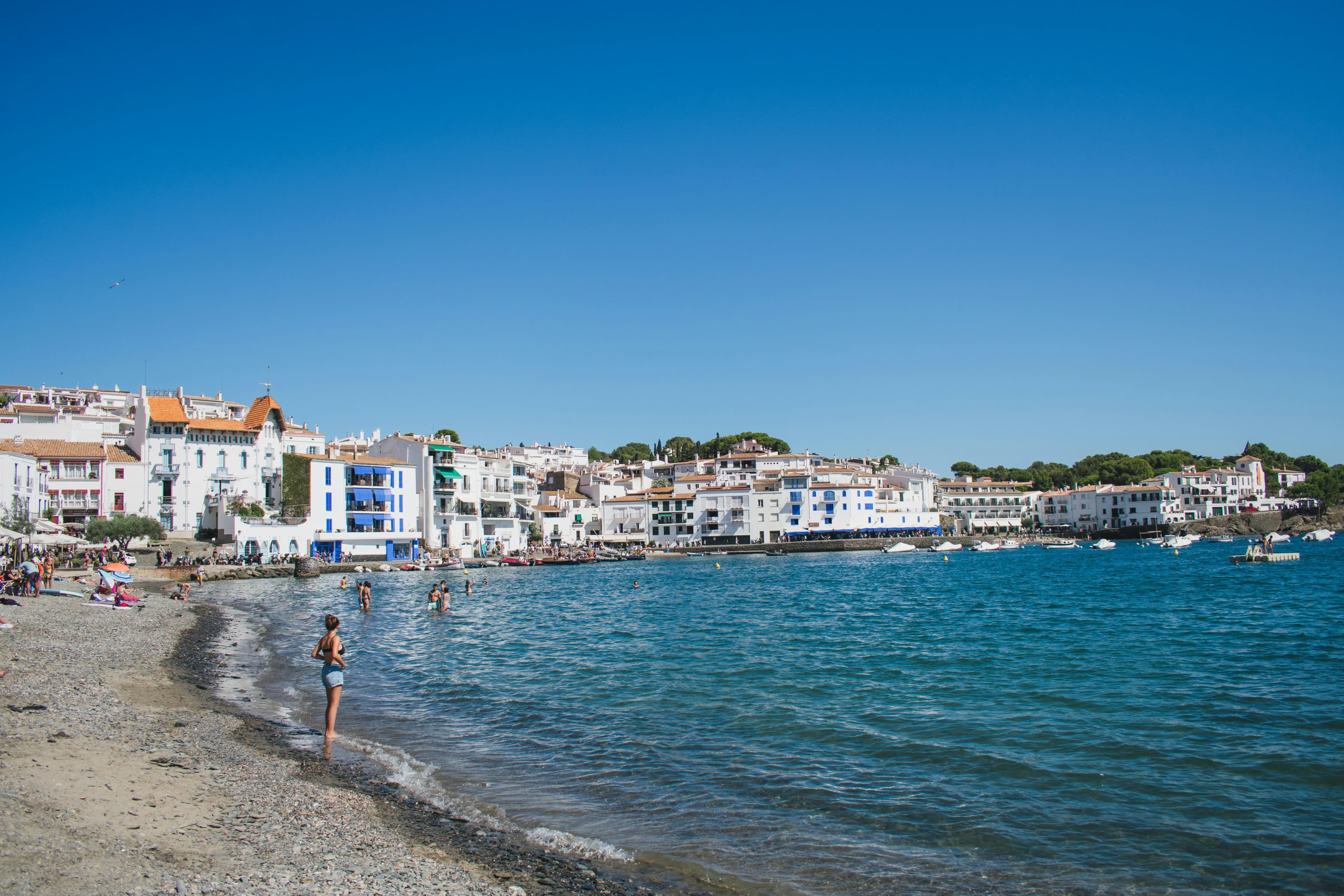The beautiful white buildings of Cadaqués are clustered around a sunny beach, with people swimming and relaxing on the shore.