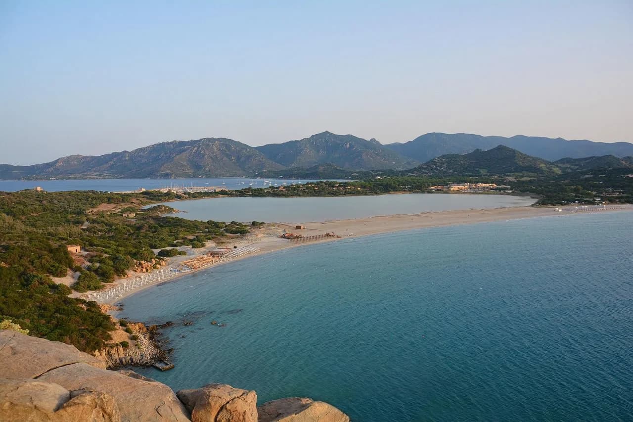 An aerial view of the coastline shows a long, sandy beach and a wide bay, with a large resort and green hills in the distance.