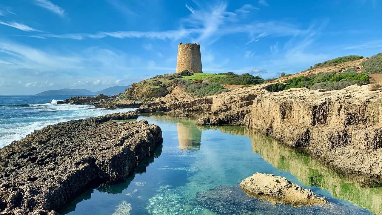 A historic stone tower stands on a rocky hill overlooking a tranquil, shallow pool of water by the sea.