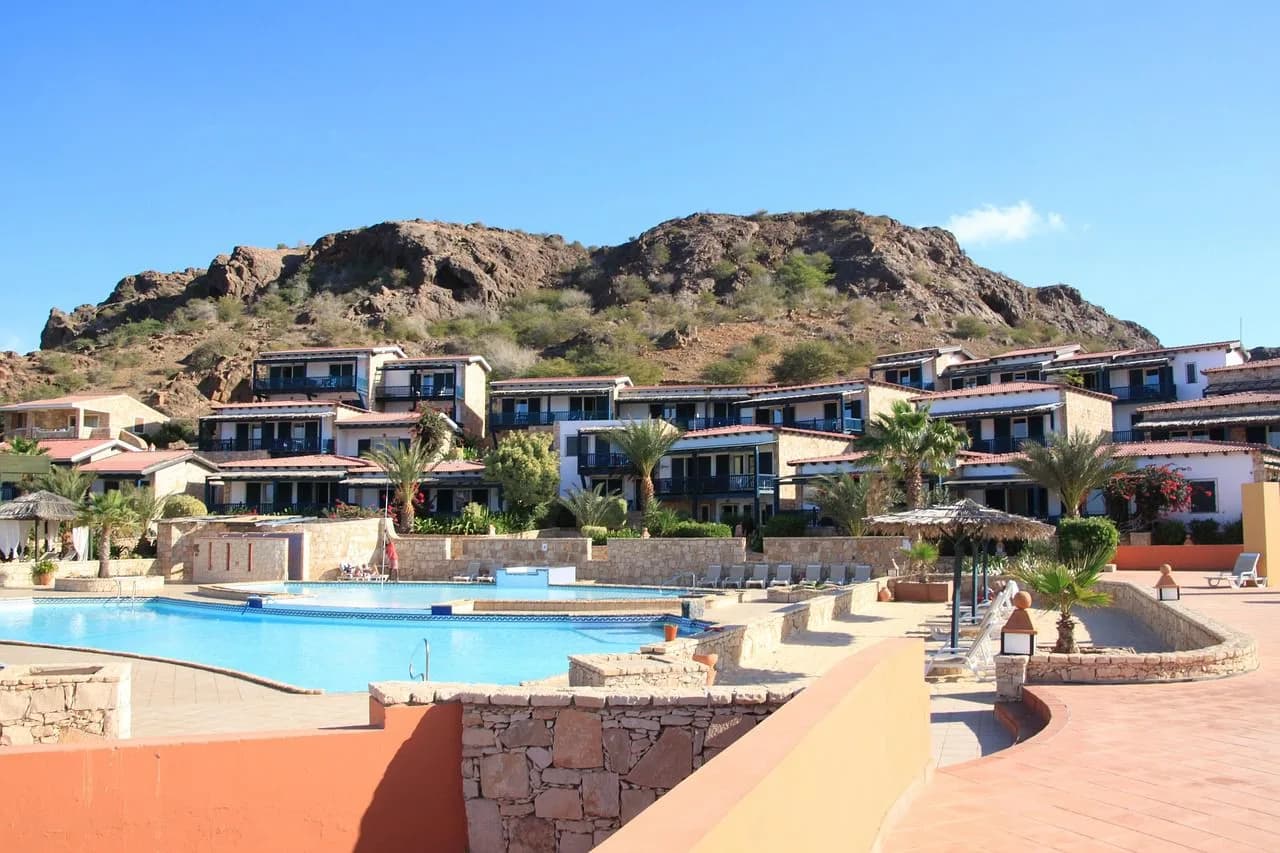 A tranquil resort pool is surrounded by palm trees and modern buildings, with rocky hills in the background.