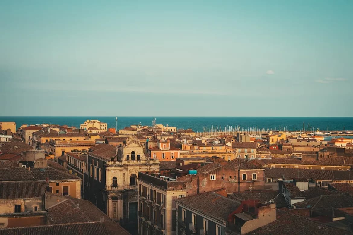 An aerial view shows the dense rooftops of Catania's historic buildings stretching toward the sea, with sailboats visible in the harbor.