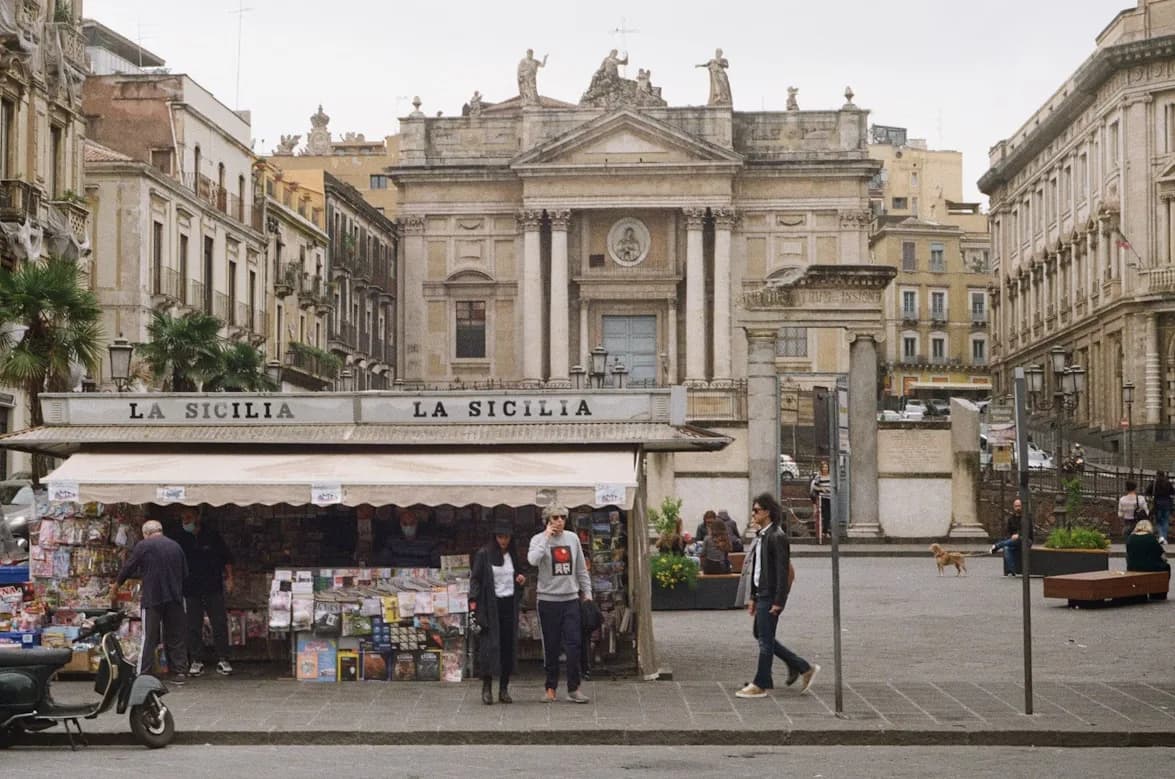A traditional market stall with a wide variety of newspapers and magazines is set up in a city square, surrounded by historic buildings.