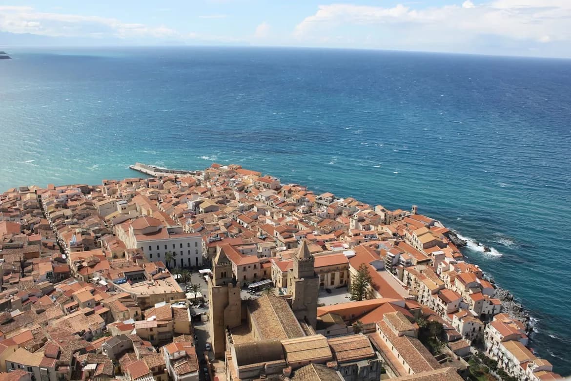 A high-angle view captures the dense cluster of red-roofed buildings in Cefalù, with the sea stretching to the horizon.