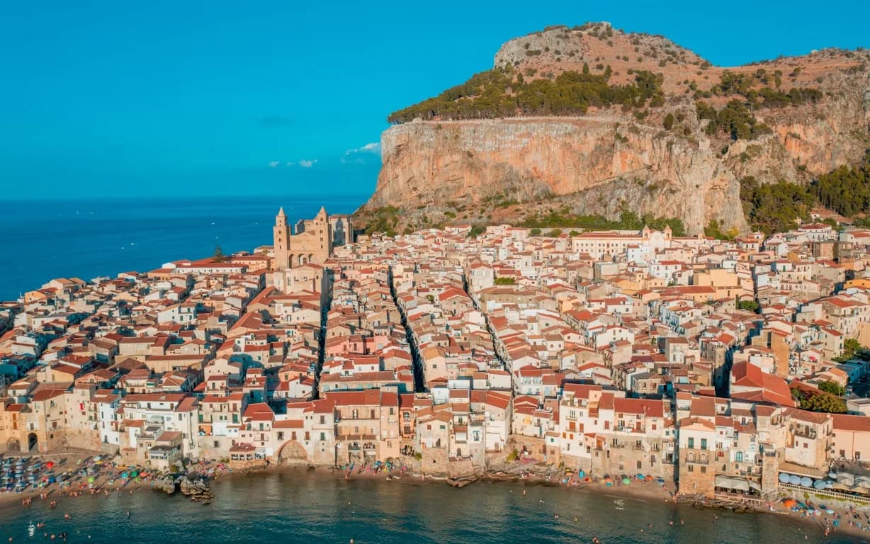 An aerial view captures the charming town of Cefalù, with its cluster of red-roofed buildings nestled against the sea and the dramatic La Rocca cliff.