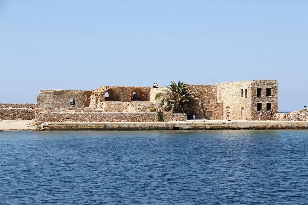 The ruins of a large, stone structure with a palm tree at its center sit on the edge of the water.