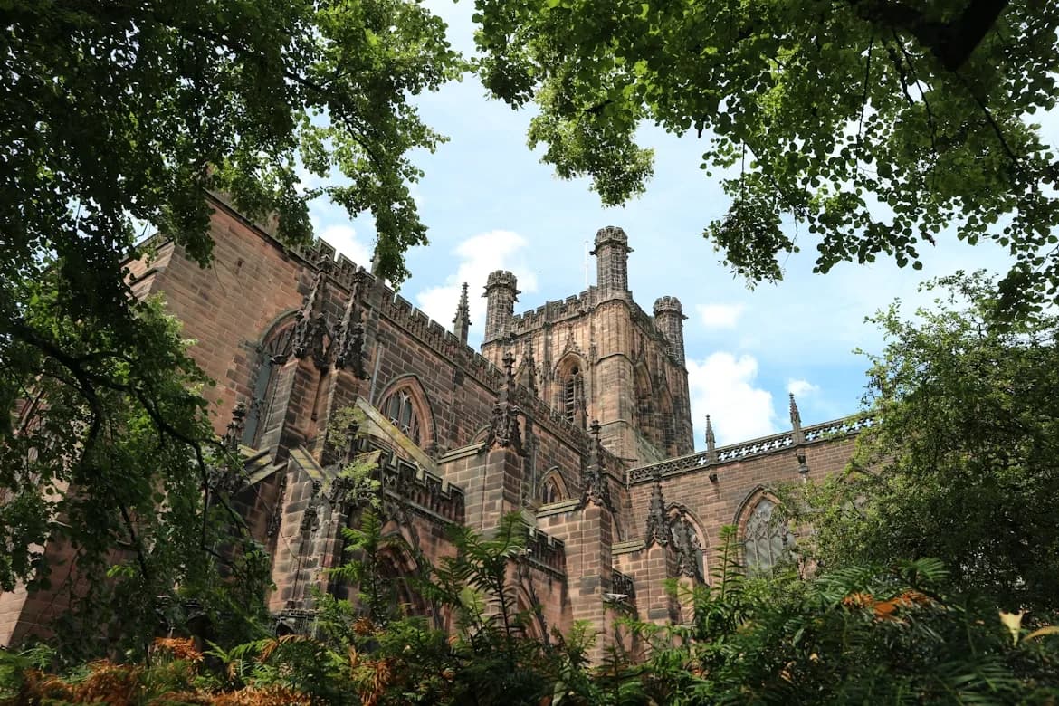 The red sandstone walls and gothic architecture of Chester Cathedral are framed by lush green trees and foliage.
