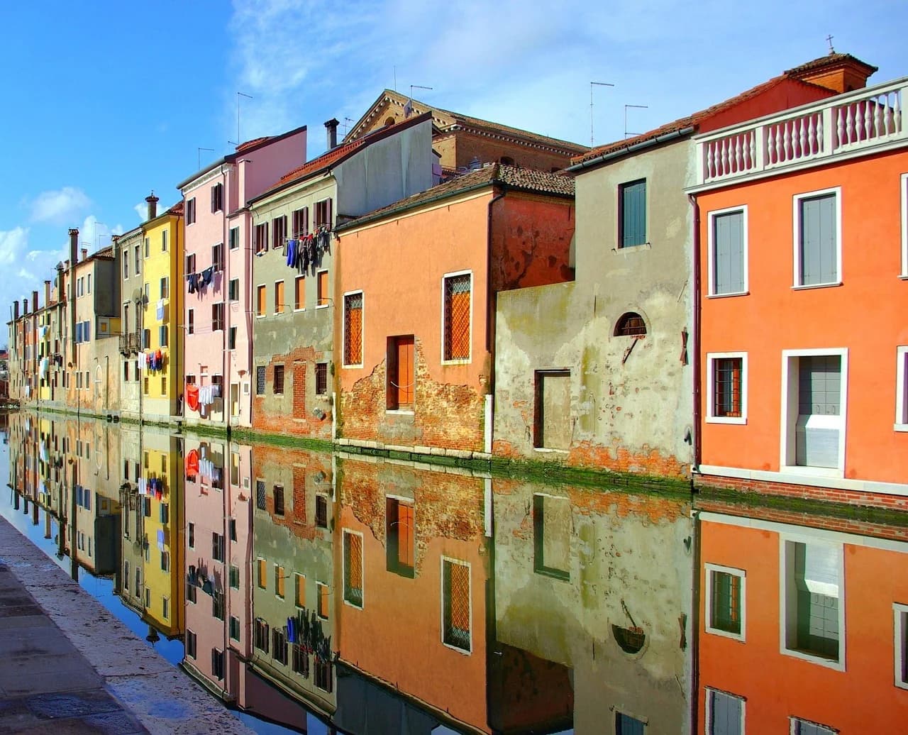 Colorful traditional houses are reflected in the tranquil waters of a canal, with a bright blue sky overhead.
