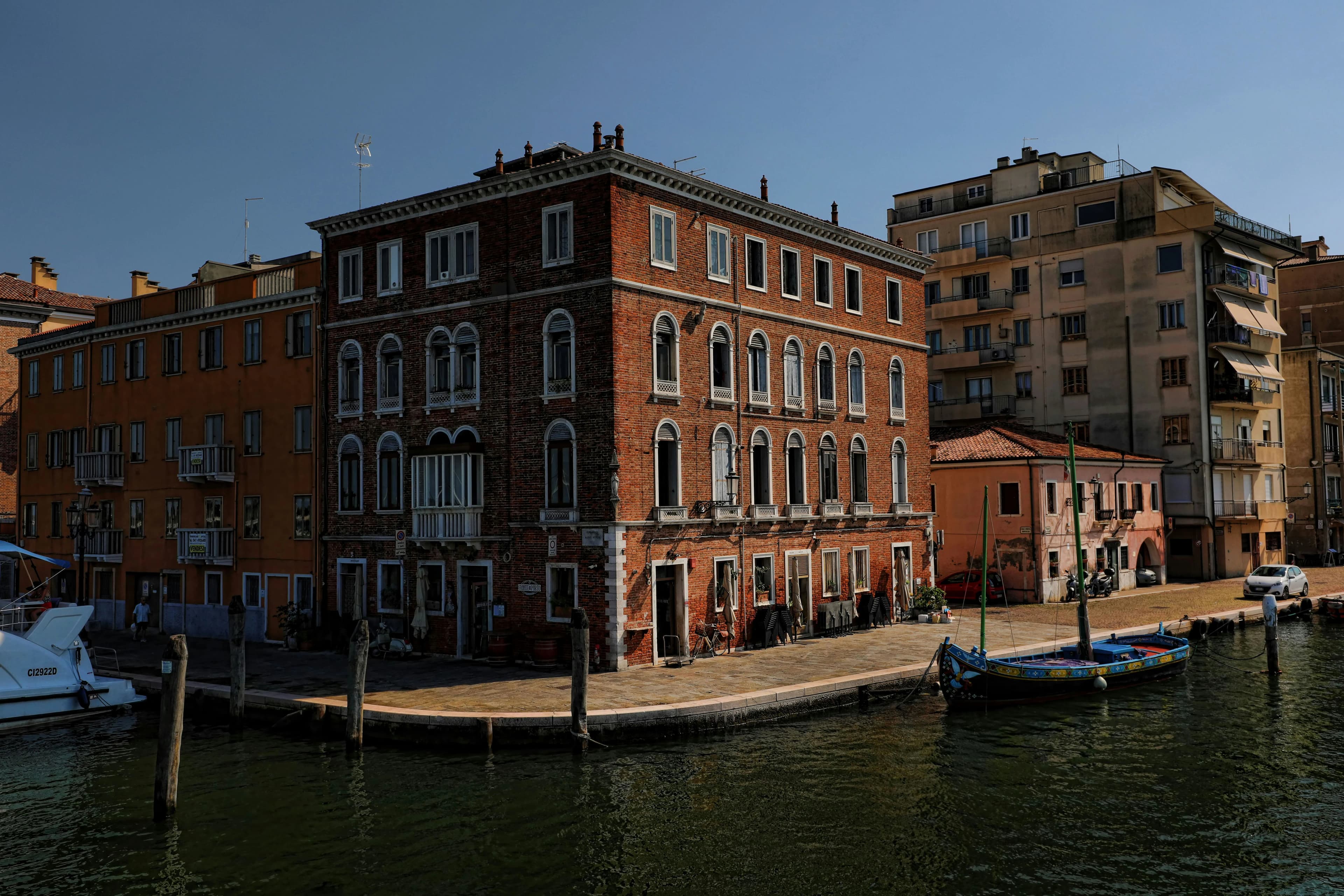 A traditional brick building with unique architectural details sits at the edge of a canal, with a small boat moored nearby.