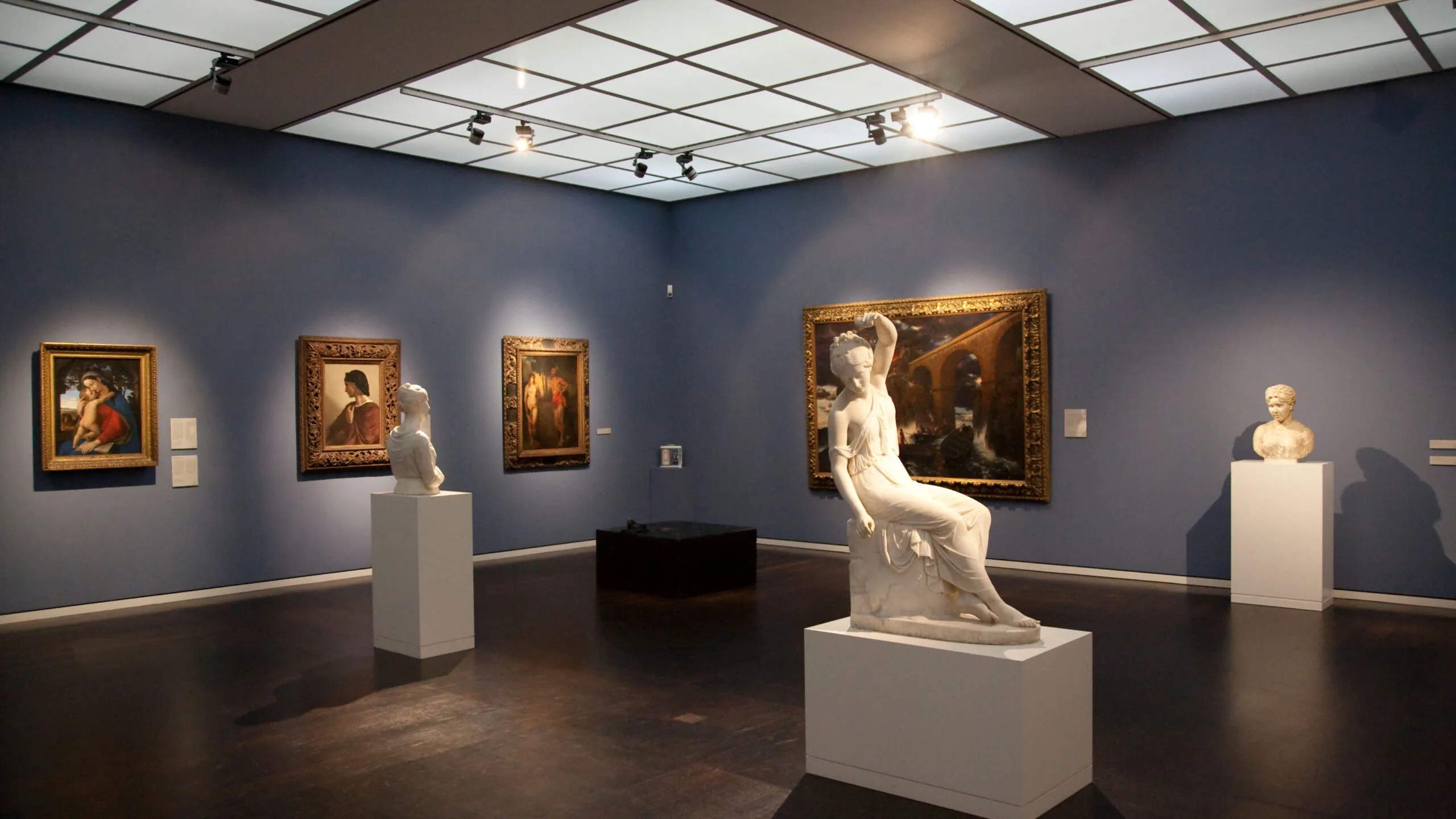 A gallery room with dark blue walls showcases a collection of classical sculptures and paintings, highlighting the city's rich art scene.