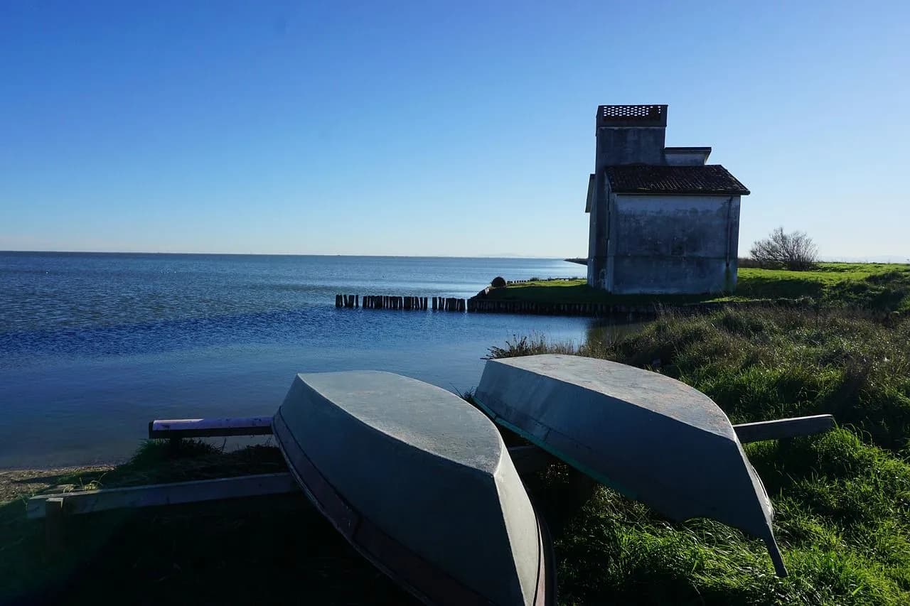 Two small boats are resting on the shore next to a weathered building on a tranquil lake.
