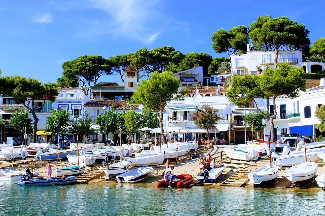 Small boats are pulled up on a sunny beach, with charming white buildings and lush trees in the background.