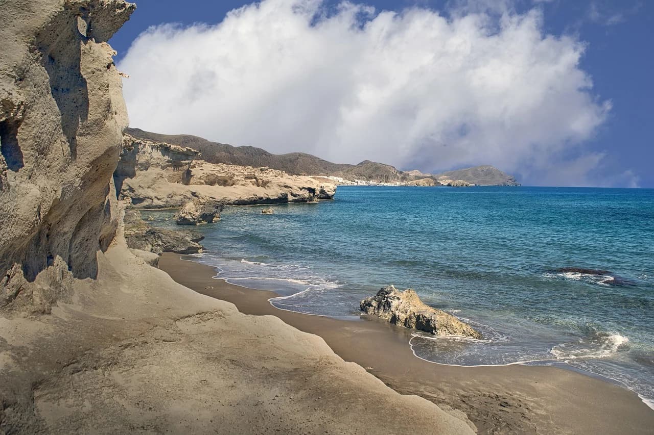 A secluded sandy beach with impressive rock formations is set against the clear blue sea and a cloudy sky.