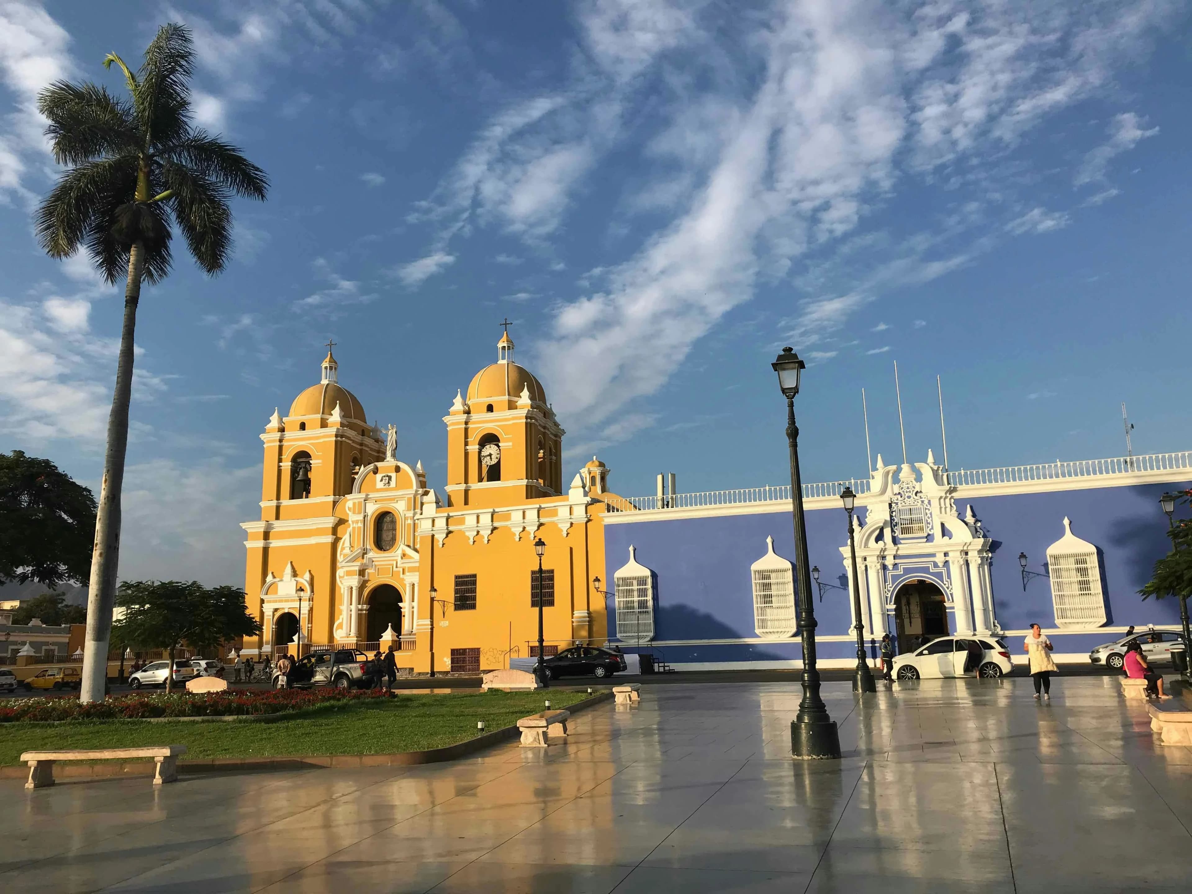 A historic, yellow and blue church with twin bell towers stands in a sun-drenched town square, with a palm tree on the left.