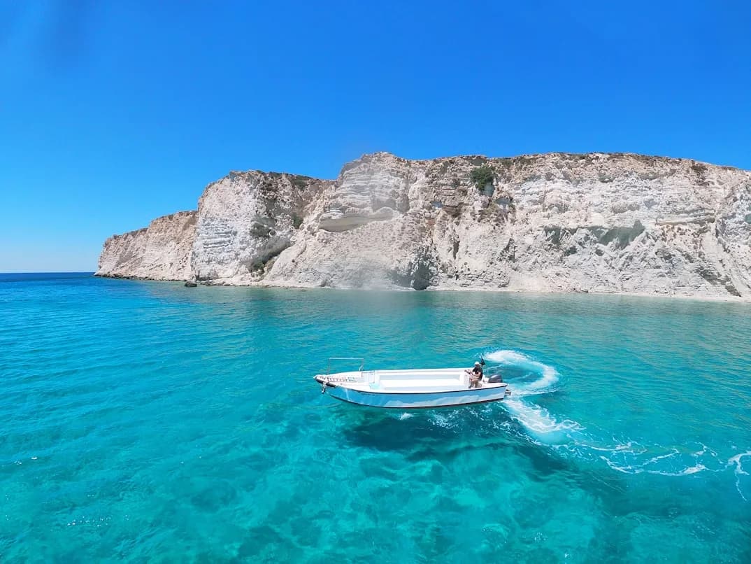 A small white boat with a motor zips through the clear turquoise waters, with towering white cliffs in the background.