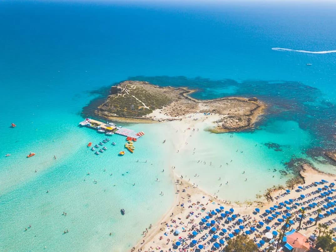 An aerial view of a peninsula with a sandy beach and a small island is surrounded by the crystal-clear waters of the Mediterranean.