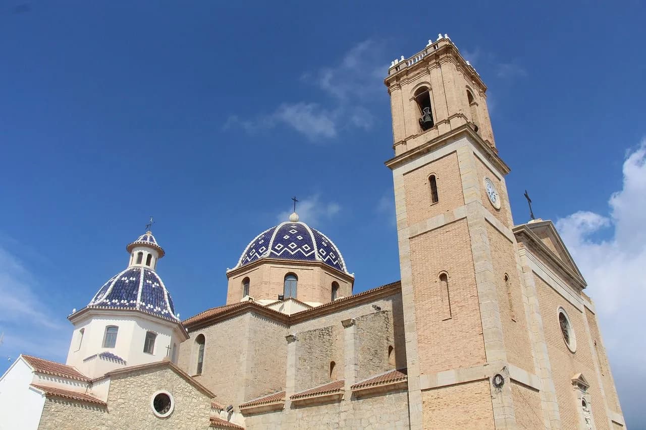 The majestic domes of a historic church with a blue and white tiled pattern are viewed from below, rising into the blue sky.