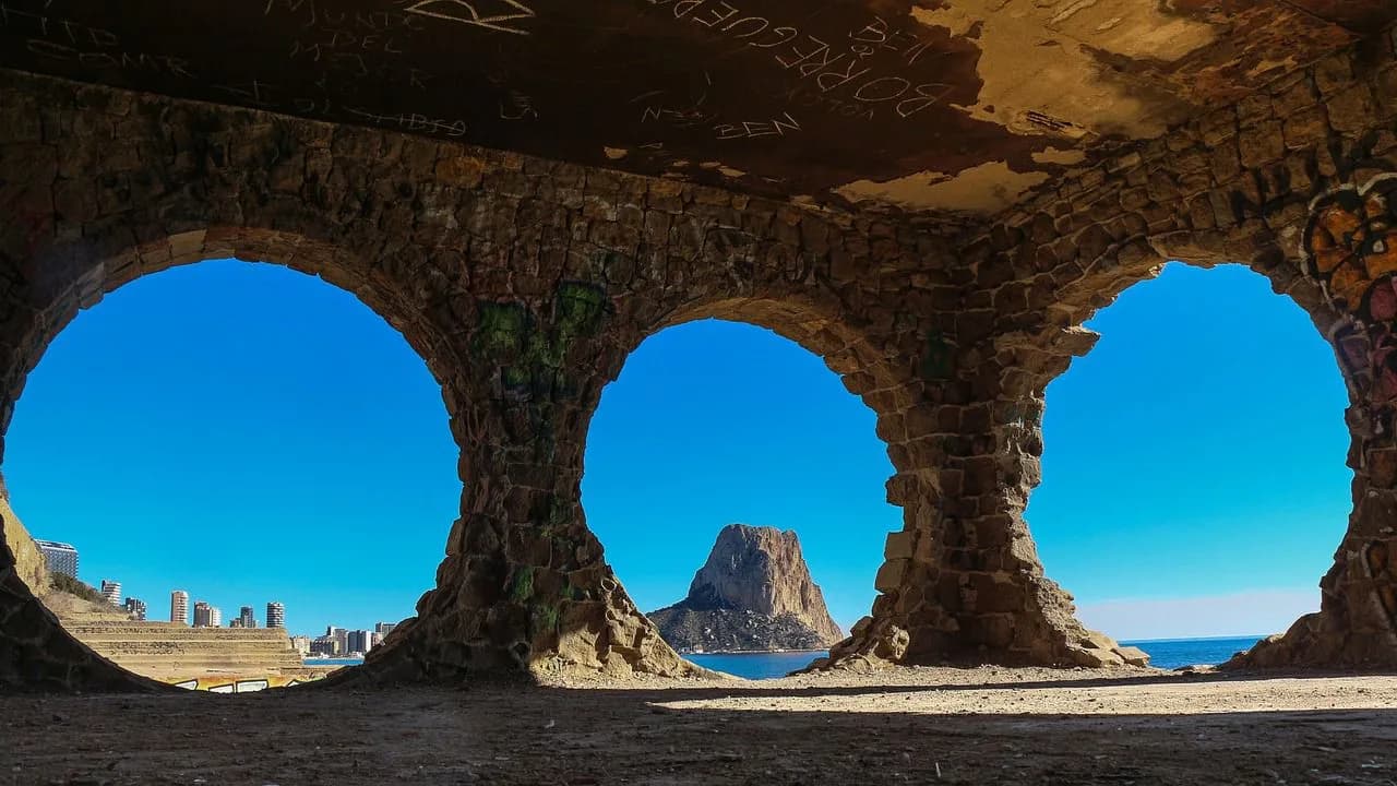A view from under a series of stone arches reveals a dramatic, rocky island in the middle of the sea.