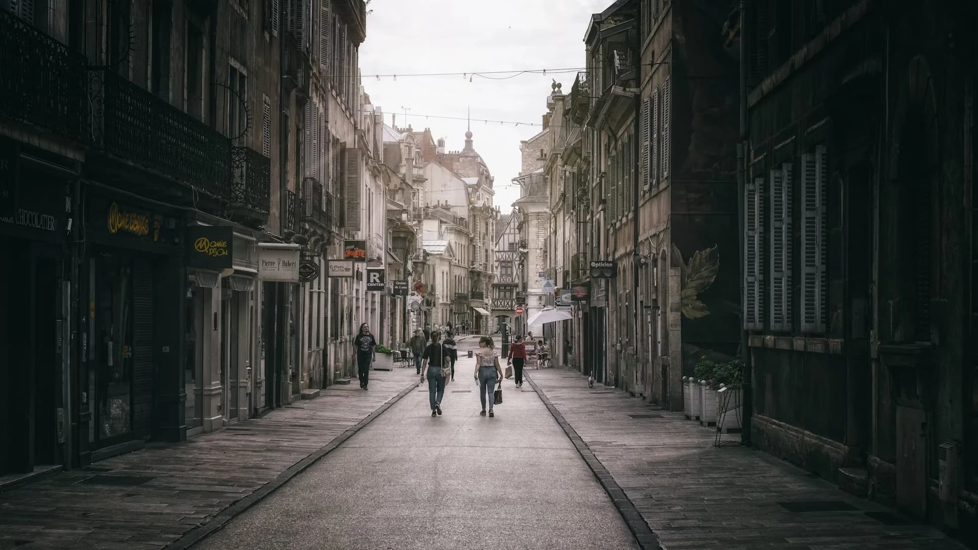 A narrow, historic street in Dijon is lined with traditional buildings and small shops, with a few people walking in the distance.
