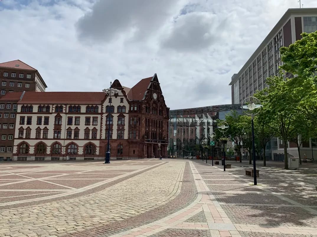 A peaceful, empty square is bordered by modern and historic buildings under a cloudy sky.