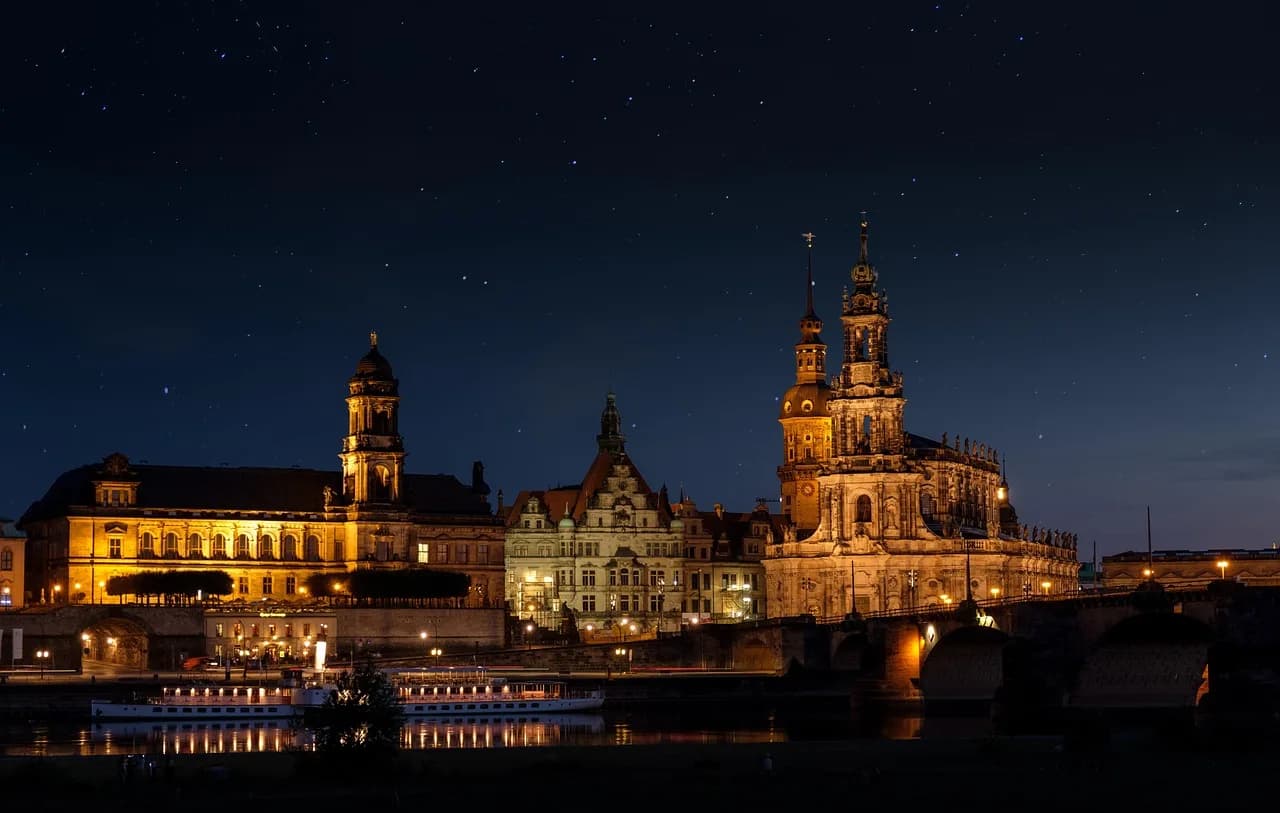 The illuminated towers of Dresden's historic skyline, including the Frauenkirche and the Hofkirche, are reflected on the tranquil Elbe River at night.