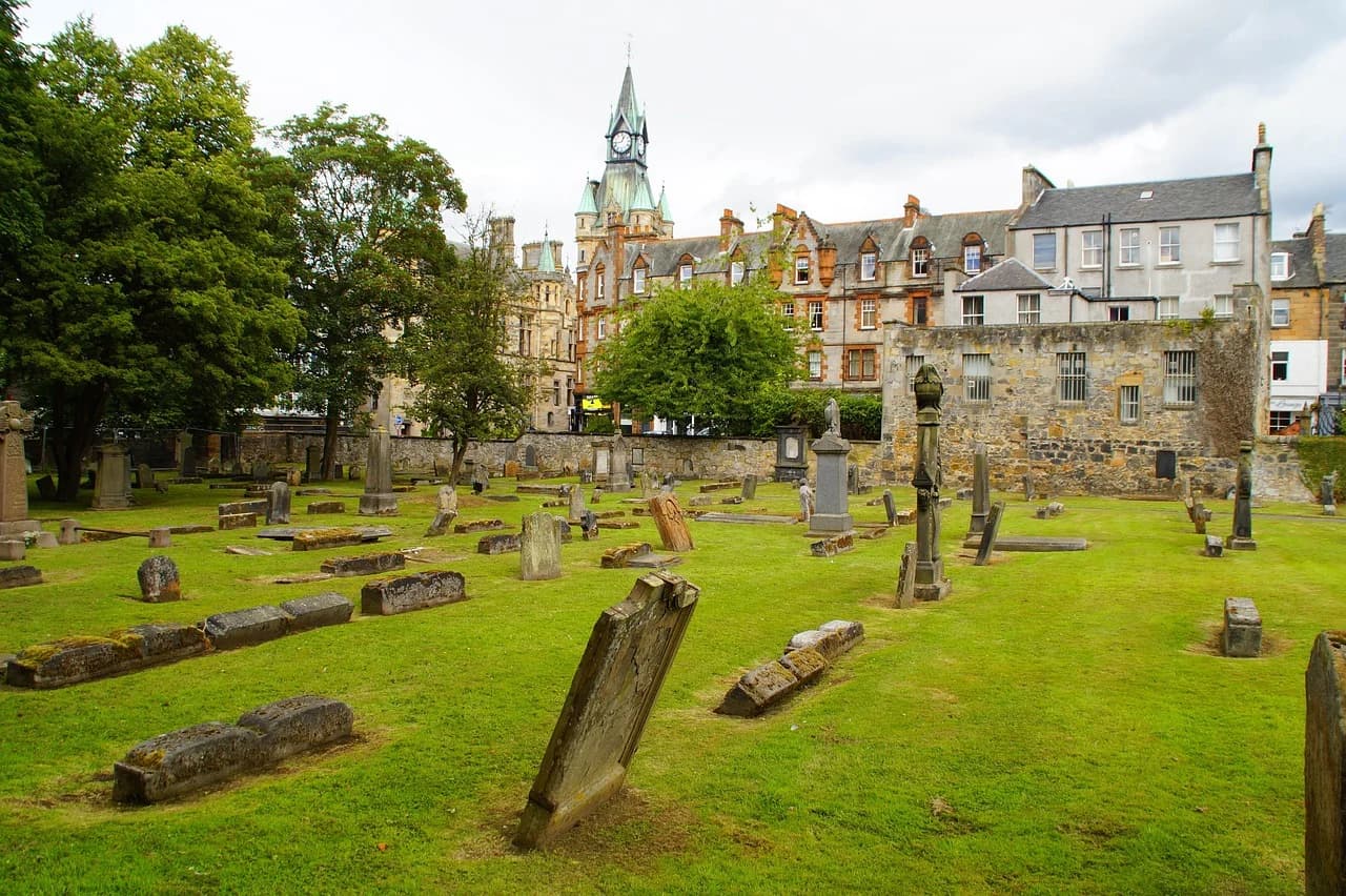 A tranquil cemetery with ancient headstones and green grass is nestled next to historic buildings with a prominent clock tower.