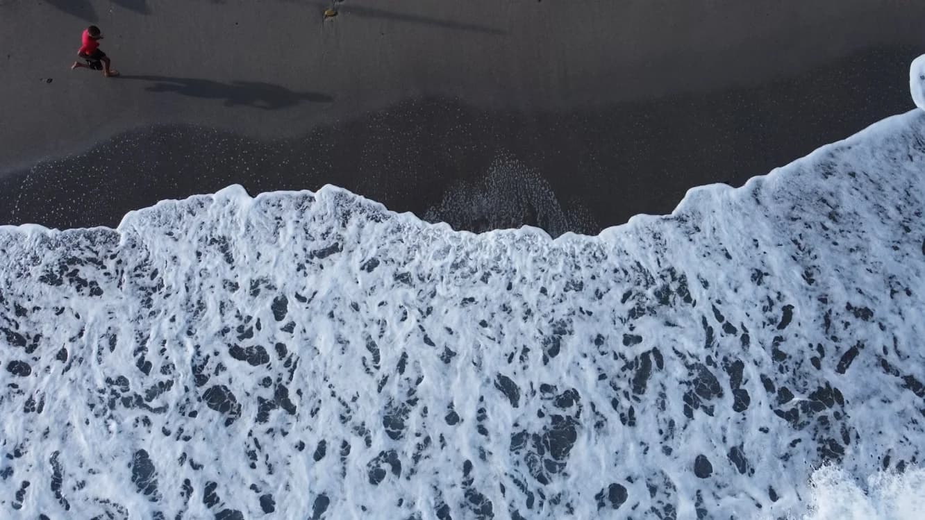 A person walks along the shore of a dark sandy beach as foamy waves crash onto the coast.