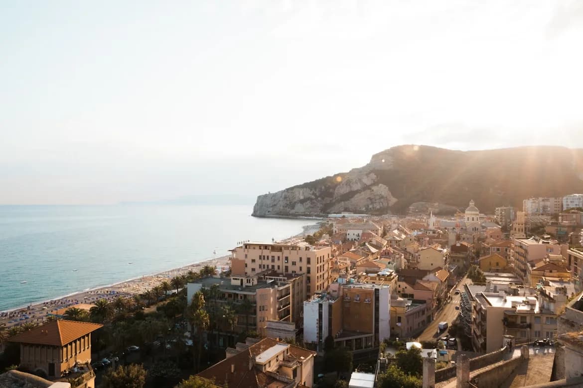 A high-angle view captures the scenic town of Finale Ligure, with its long sandy beach and beautiful buildings nestled against the hillside.