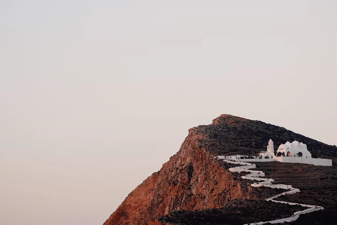 The iconic white church of Panagia stands atop a steep, rocky hill, with a zigzagging pathway leading to it.