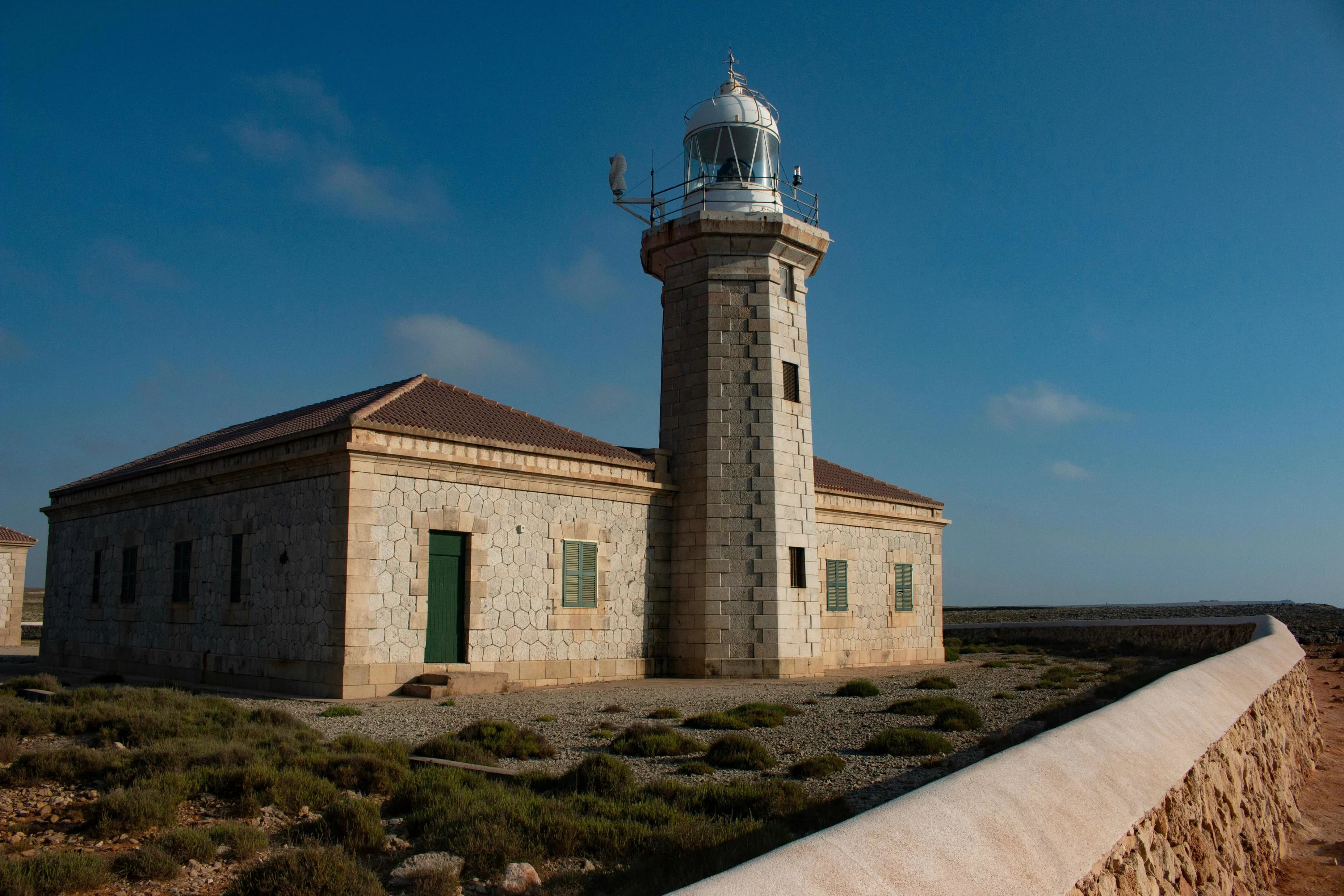 The Far de la Mola, a historic lighthouse, stands on a flat, rocky landscape, with a bright blue sky in the background.
