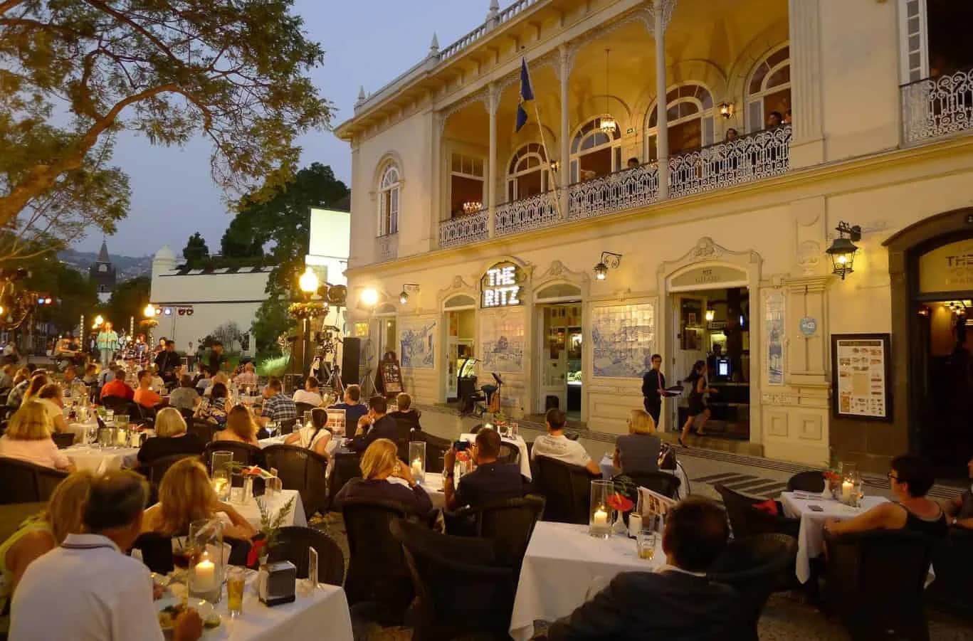 People dine at outdoor tables in front of a grand, colonial-style hotel building, with a live musician performing in the foreground.