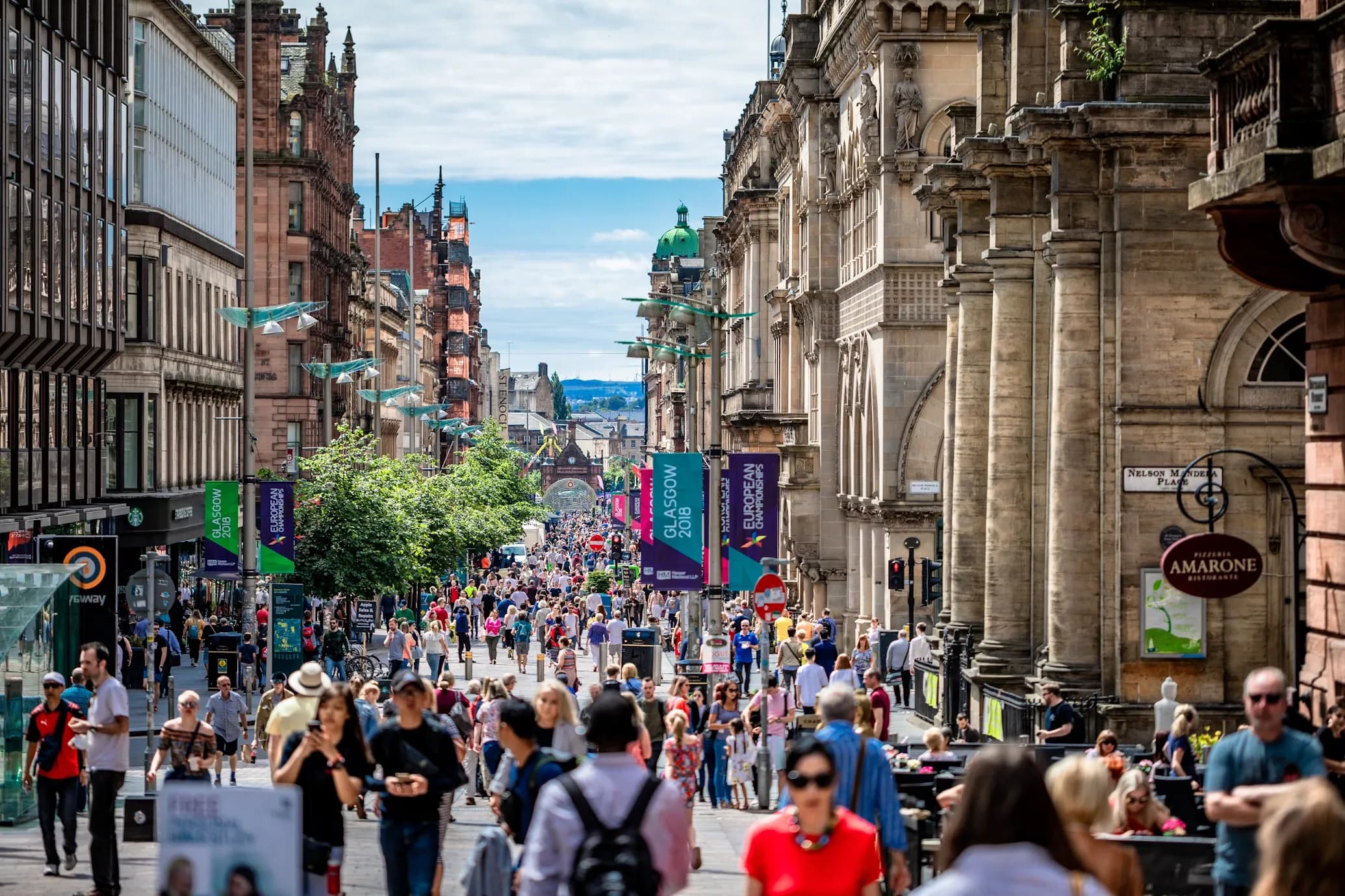 A busy city street in Glasgow is bustling with people and is lined with elegant, traditional buildings.