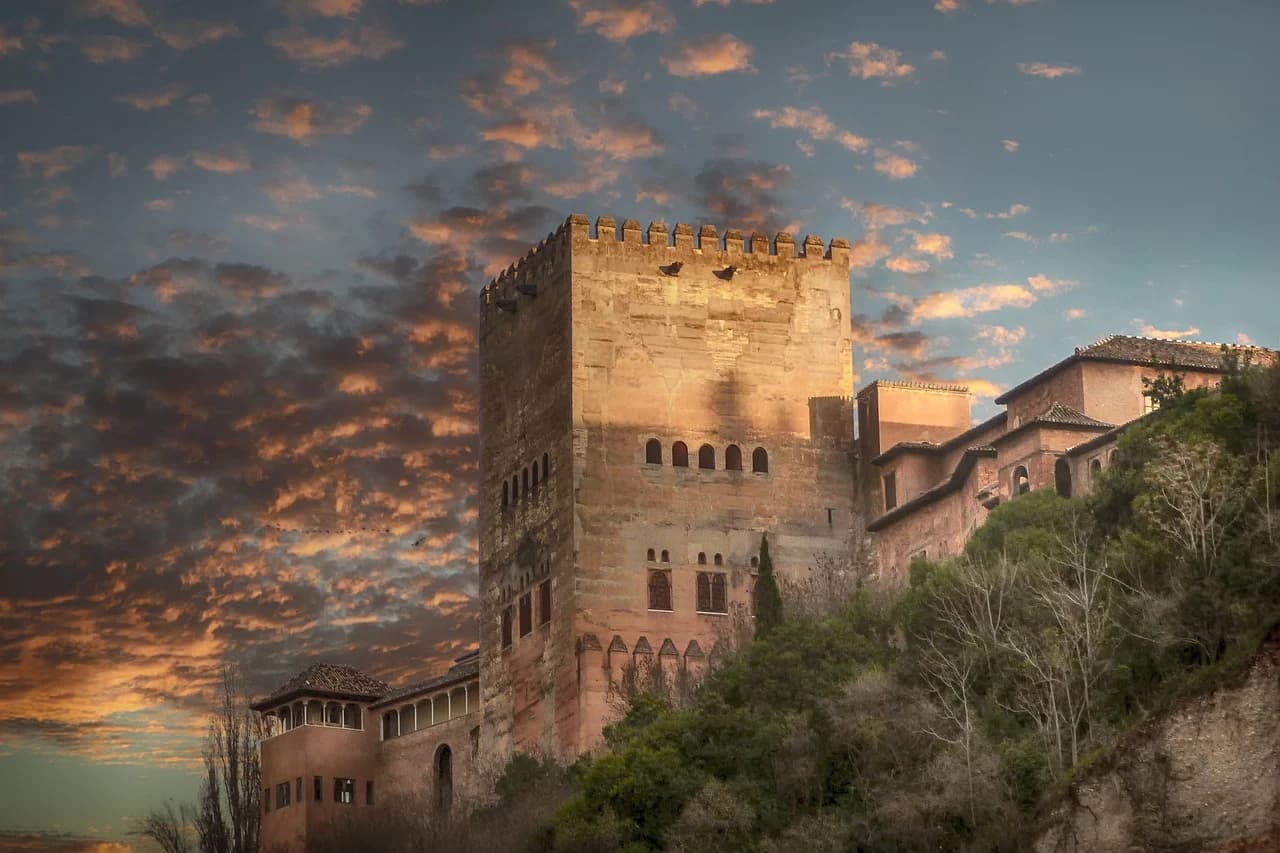 A dramatic view of the Alhambra's towers and walls is silhouetted against a vibrant, cloudy sunset.