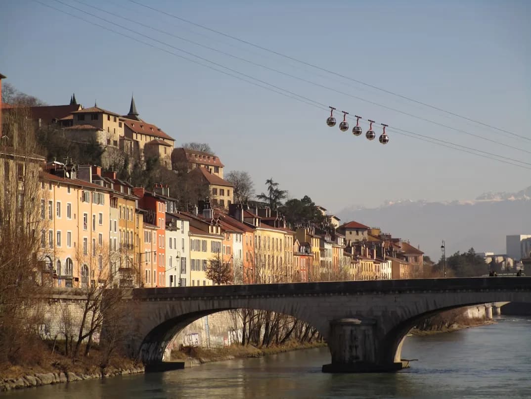 The "Bubbles" of Grenoble's cable car system glide over the Isère River, with historic buildings and the Bastille fortress on the hillside.
