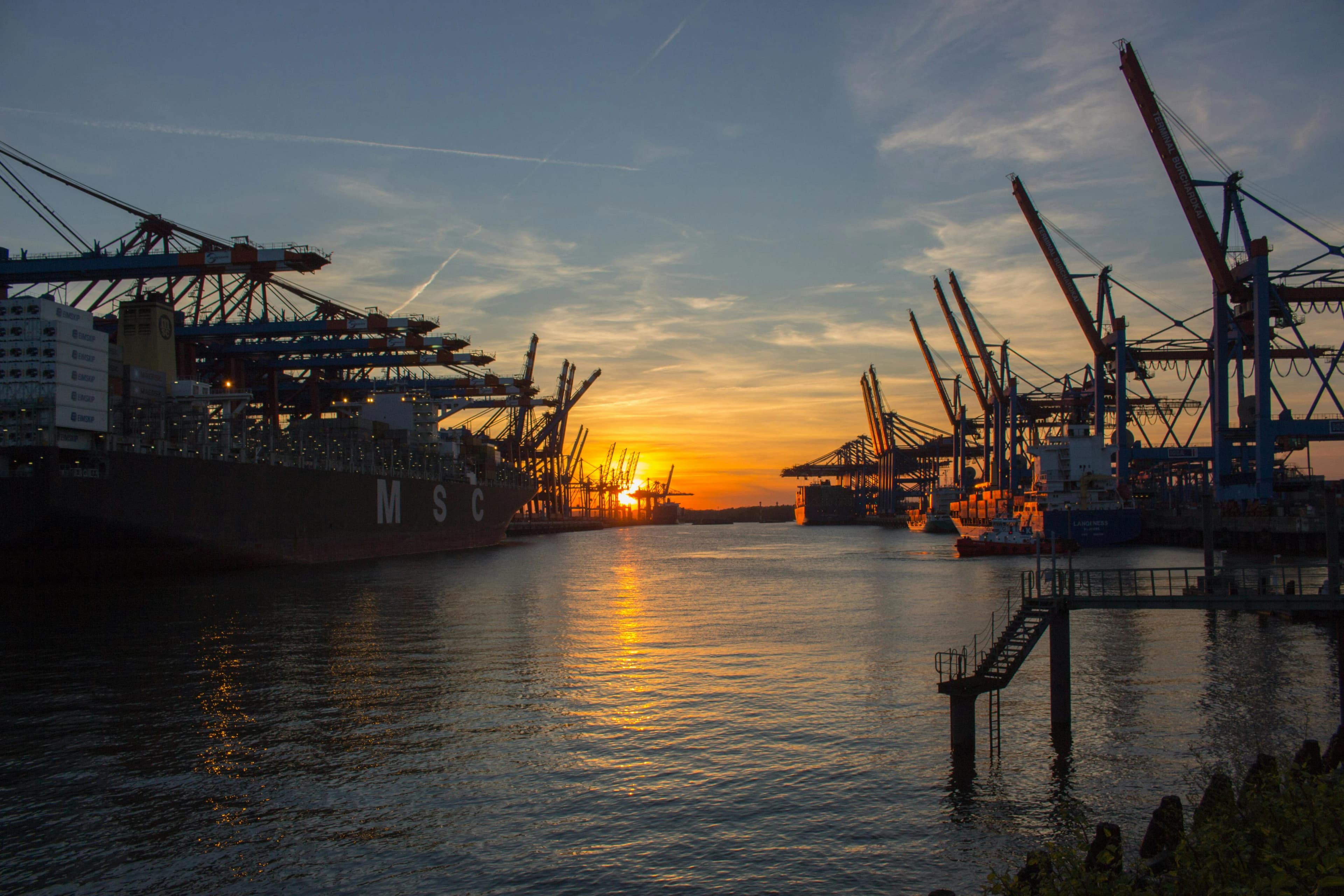 The busy port of Hamburg is bathed in the warm light of a sunset, with massive container ships and cranes silhouetted against the sky.